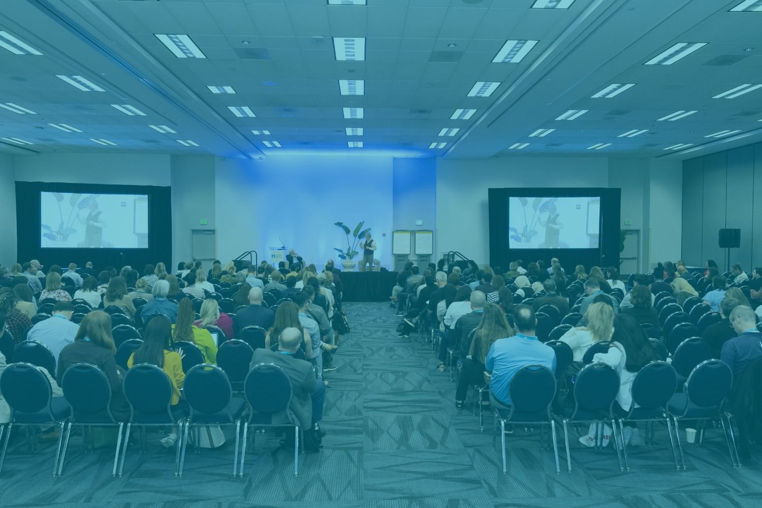 Forum 2025 Attendees Sitting in on a Session Where a Speaker is on Stage with 2 Projector Screens