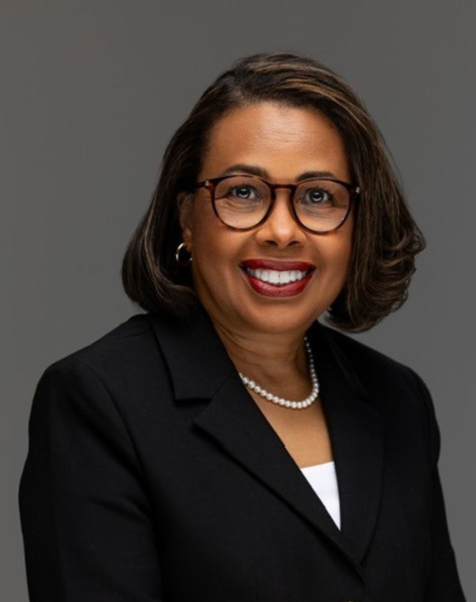 A headshot image of Sylvia Trent-Adams wearing a black blazer, glasses, and a white necklace against a gray background.